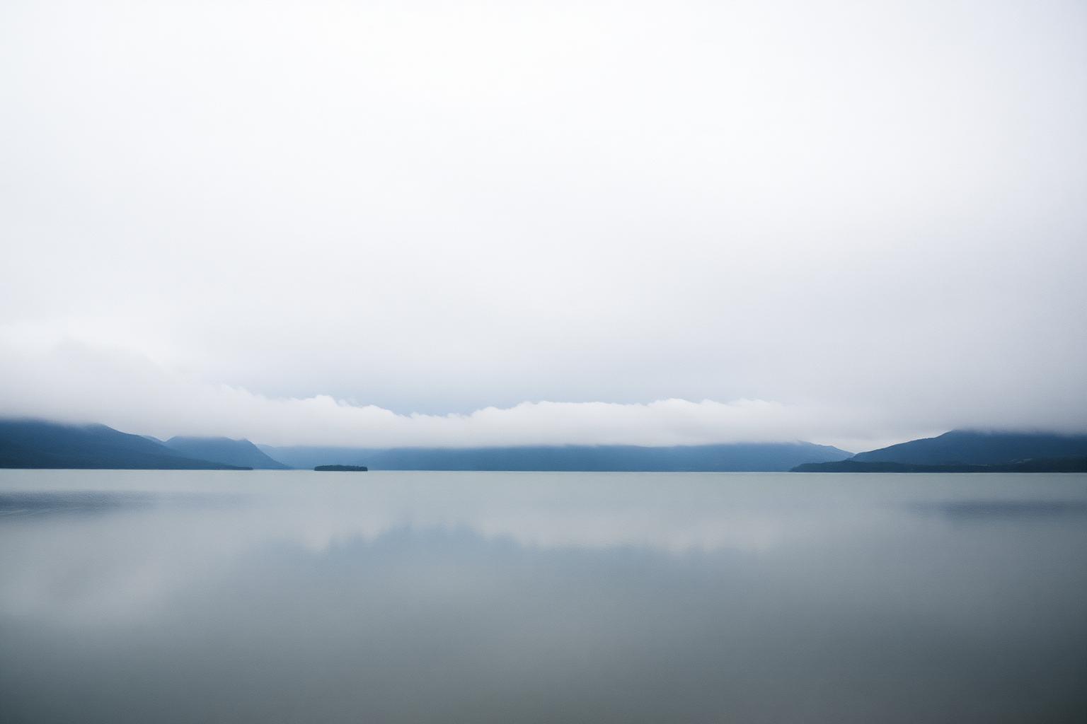 Clouds clearing over a still lake, half the sky grey and half turning blue.