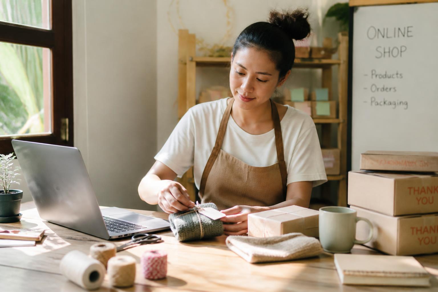 A person packing handmade products beside a laptop at a sunlit table while running a small home-based online shop.